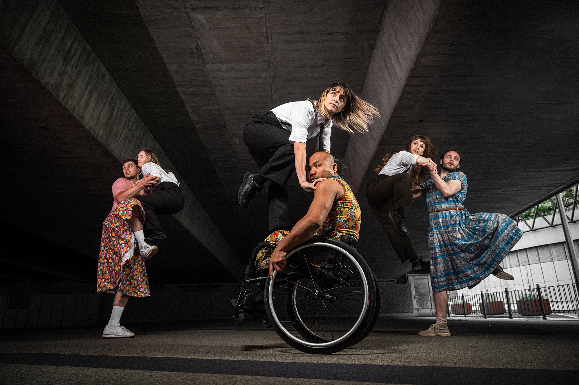 Stopgap dance company perform 'Frock' under a bridge. A black male dancer in a wheelchair is centre, lifting a female dancer. 