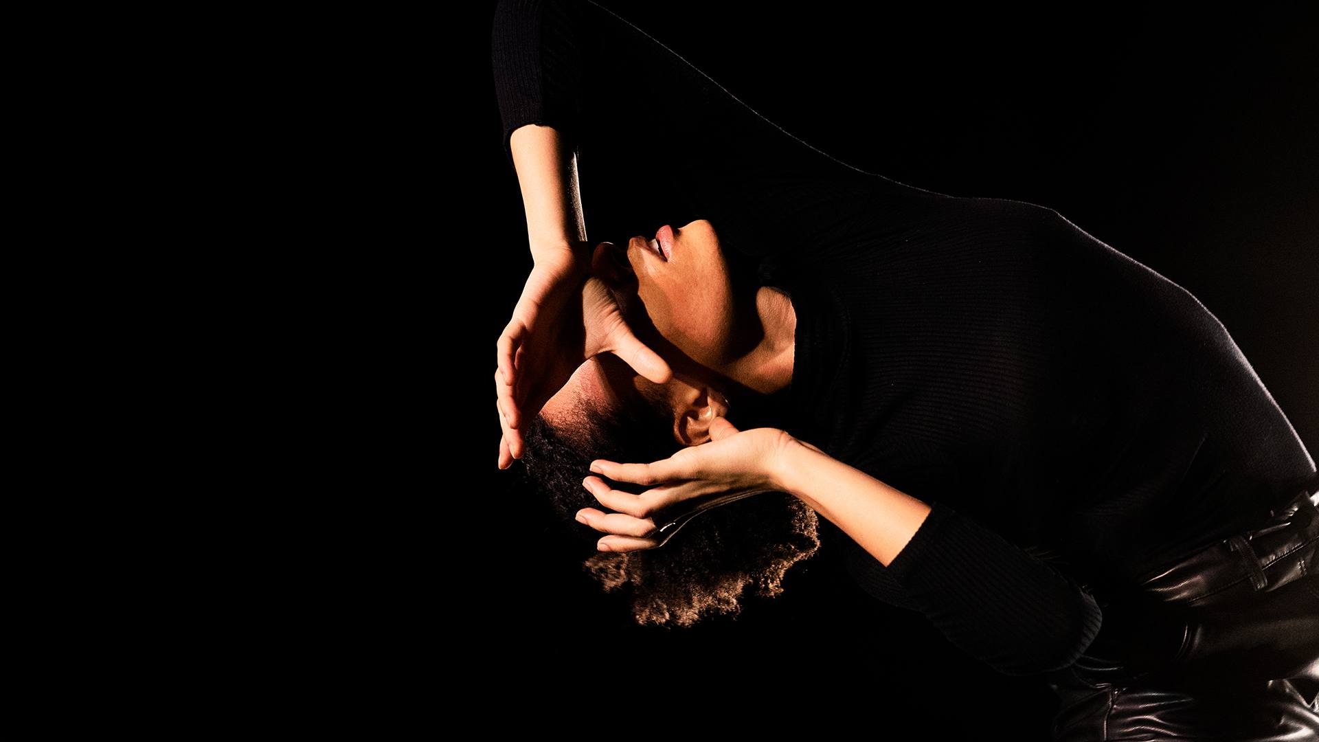 A black female dancer dressed all in black leans back with her hands grasping something imaginary