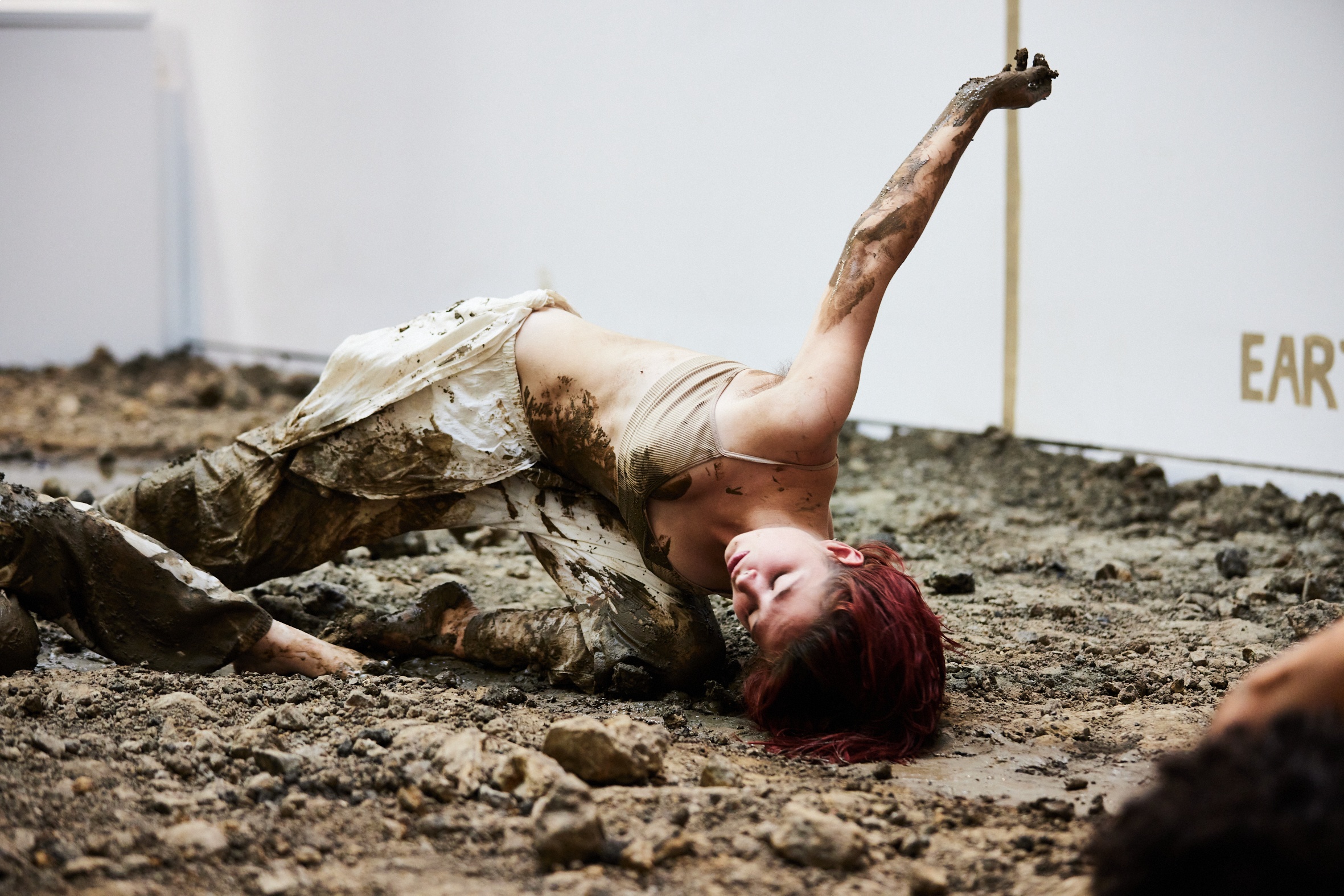 A white female dancer with red hair performs in a white gallery space on a bed of mud and rocky landscape, an installation by an artist. Her clothes and hands are covered in mud
