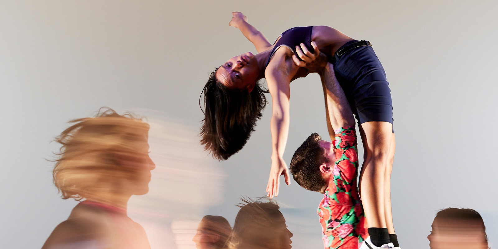 A female dancer is lifted high, arching back as she looks directly at the camera. Blurred figures run around her. 