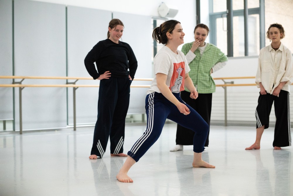 Bar Groisman in the studio with her company of dancers. Bar demonstrates a move with a smile on her face while the dancers look on excitedly