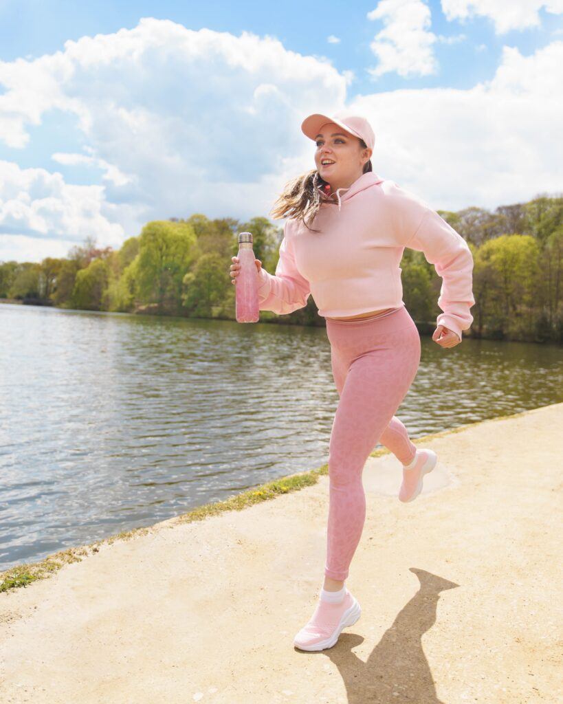 Promotional shot of Lydia Cottrell for 'Brightside'. Lydia wears baby pink running gear complete with pink cap and pink water bottle and trainers, and is captured mid-run by a lake in a park. 